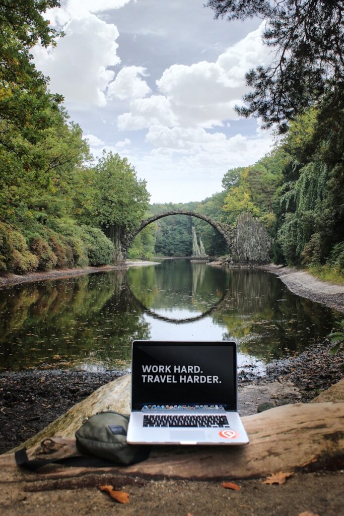 silver MacBook Pro on brown wood log looking out at body of water. The words on the screen read 'work hard, travel harder'