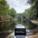 silver MacBook Pro on brown wood log looking out at body of water. The words on the screen read 'work hard, travel harder'